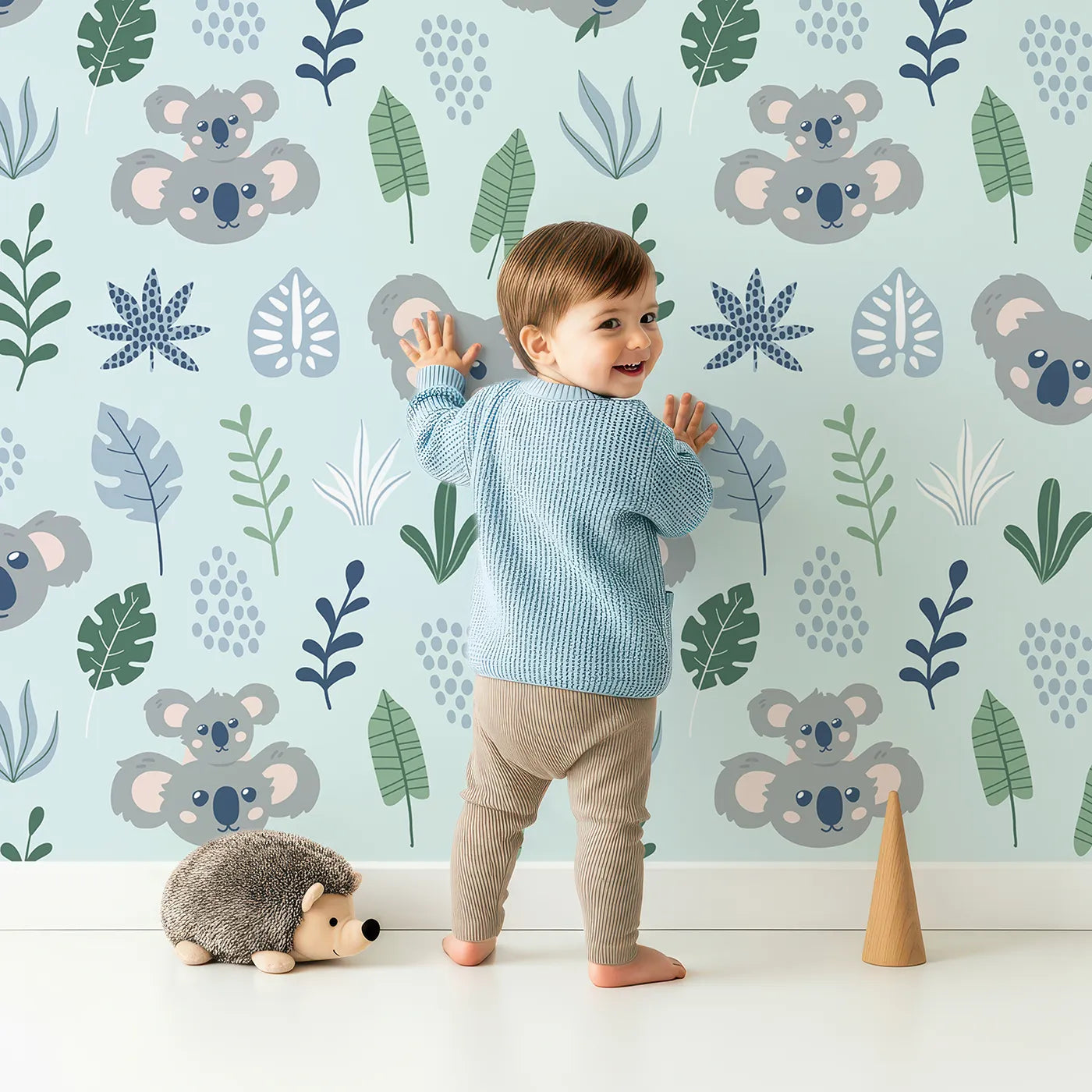 Lovely child standing in front of Whimsy Tots Koala Cuddle Grove Wallpaper with koala, leaf, jungle, botanical, animal in design.