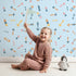 Lovely child standing in front of Whimsy Tots Boho Arrows Wallpaper in Blue with arrow, triangle, dot in design. Lovely child standing in front of Whimsy Tots Boho Arrows Wallpaper in Blue with arrow, triangle, dot in design.