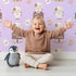 Lovely child standing in front of Whimsy Tots Moo Farm Friends Wallpaper in Purple with cow, bottle, bell, wheat, sign in design. Lovely child standing in front of Whimsy Tots Moo Farm Friends Wallpaper in Purple with cow, bottle, bell, wheat, sign in design.