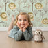 Lovely child standing in front of Whimsy Tots Little Lion Nap Wallpaper in Green with lion, tree, footprint in design. Lovely child standing in front of Whimsy Tots Little Lion Nap Wallpaper in Green with lion, tree, footprint in design.