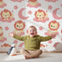 Lovely child standing in front of Whimsy Tots Starry Safari Dreams Wallpaper in Pink with lion, cloud, moon, star in design. Lovely child standing in front of Whimsy Tots Starry Safari Dreams Wallpaper in Pink with lion, cloud, moon, star in design.