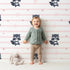 Lovely child standing in front of Whimsy Tots Starlit Raccoon Stripes Wallpaper in Pink with raccoon, star, stripe in design. Lovely child standing in front of Whimsy Tots Starlit Raccoon Stripes Wallpaper in Pink with raccoon, star, stripe in design.