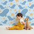 Lovely child standing in front of Whimsy Tots Starry Whale Friends Wallpaper in Blue with whale, starfish, bubble, dot in design. Lovely child standing in front of Whimsy Tots Starry Whale Friends Wallpaper in Blue with whale, starfish, bubble, dot in design.