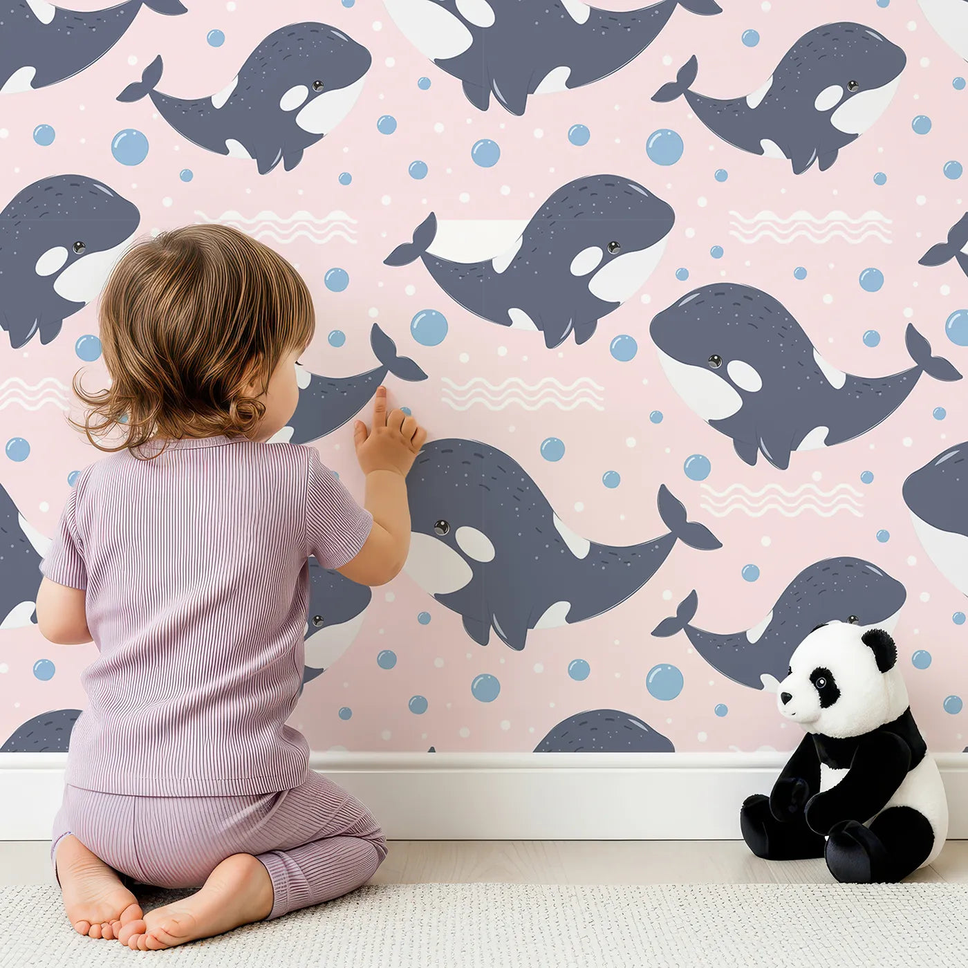 Lovely child standing in front of Whimsy Tots Orca Whale Wonder Wallpaper in Blush Pink with whale, orca, bubble, wave in design.