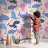 Lovely child standing in front of Whimsy Tots Ocean Buddy Parade Wallpaper in Blush Pink with whale, dolphin, shark, narwhal, octopus, starfish, bubble in design. Lovely child standing in front of Whimsy Tots Ocean Buddy Parade Wallpaper in Blush Pink with whale, dolphin, shark, narwhal, octopus, starfish, bubble in design.