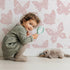 Lovely child standing in front of Whimsy Tots Celestial Wings Butterfly Wallpaper in Blush Pink with butterfly, moon, star, swirl in design. Lovely child standing in front of Whimsy Tots Celestial Wings Butterfly Wallpaper in Blush Pink with butterfly, moon, star, swirl in design.