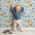 Lovely child standing in front of Whimsy Tots Meadow Cuddles Deer Wallpaper in Sky Blue with deer, flower, leaf, blossom in design. Lovely child standing in front of Whimsy Tots Meadow Cuddles Deer Wallpaper in Sky Blue with deer, flower, leaf, blossom in design.