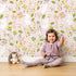 Lovely child standing in front of Whimsy Tots Bloomy Bunny Meadow Wallpaper in Petal Pink with rabbit, flower, leaf, blossom in design. Lovely child standing in front of Whimsy Tots Bloomy Bunny Meadow Wallpaper in Petal Pink with rabbit, flower, leaf, blossom in design.