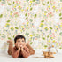 Lovely child standing in front of Whimsy Tots Bloomy Bunny Meadow Wallpaper in Oat Beige with rabbit, flower, leaf, blossom in design. Lovely child standing in front of Whimsy Tots Bloomy Bunny Meadow Wallpaper in Oat Beige with rabbit, flower, leaf, blossom in design.