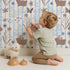 Lovely child standing in front of Whimsy Tots Meadow Hop Bunnies Wallpaper in Sky Blue with rabbit, flower, bunny, leaf, stripe in design. Lovely child standing in front of Whimsy Tots Meadow Hop Bunnies Wallpaper in Sky Blue with rabbit, flower, bunny, leaf, stripe in design.