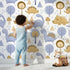 Lovely child standing in front of Whimsy Tots Jurassic Forest Friends Wallpaper in White with dinosaur, tree, triangle, dino, forest in design. Lovely child standing in front of Whimsy Tots Jurassic Forest Friends Wallpaper in White with dinosaur, tree, triangle, dino, forest in design.
