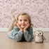 Lovely child standing in front of Whimsy Tots Desert Dino Parade Wallpaper in Blush Pink with dinosaur, cactus, flower in design. Lovely child standing in front of Whimsy Tots Desert Dino Parade Wallpaper in Blush Pink with dinosaur, cactus, flower in design.