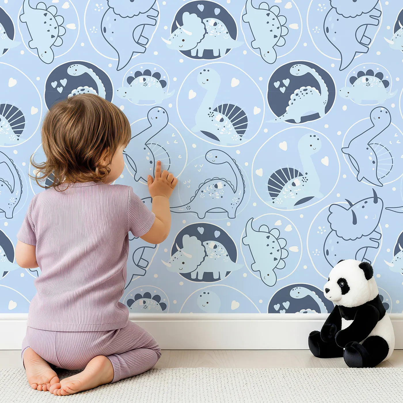 Lovely child standing in front of Whimsy Tots Dino Circle Parade Wallpaper in Sky Blue with dinosaur, flower, heart, circle in design.