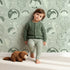 Lovely child standing in front of Whimsy Tots Dino Circle Parade Wallpaper in Sage Green with dinosaur, flower, heart, circle in design. Lovely child standing in front of Whimsy Tots Dino Circle Parade Wallpaper in Sage Green with dinosaur, flower, heart, circle in design.