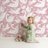 Lovely child standing in front of Whimsy Tots Swan Princess Waltz Wallpaper in Blush Pink with swan, crown, wing, heart, star in design. Lovely child standing in front of Whimsy Tots Swan Princess Waltz Wallpaper in Blush Pink with swan, crown, wing, heart, star in design.