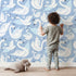 Lovely child standing in front of Whimsy Tots Swan Princess Waltz Wallpaper in Blue with swan, crown, wing, heart, star in design. Lovely child standing in front of Whimsy Tots Swan Princess Waltz Wallpaper in Blue with swan, crown, wing, heart, star in design.