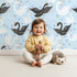 Lovely child standing in front of Whimsy Tots Royal Swan Parade Wallpaper in Sky Blue with swan, crown, heart, star, wing in design. Lovely child standing in front of Whimsy Tots Royal Swan Parade Wallpaper in Sky Blue with swan, crown, heart, star, wing in design.