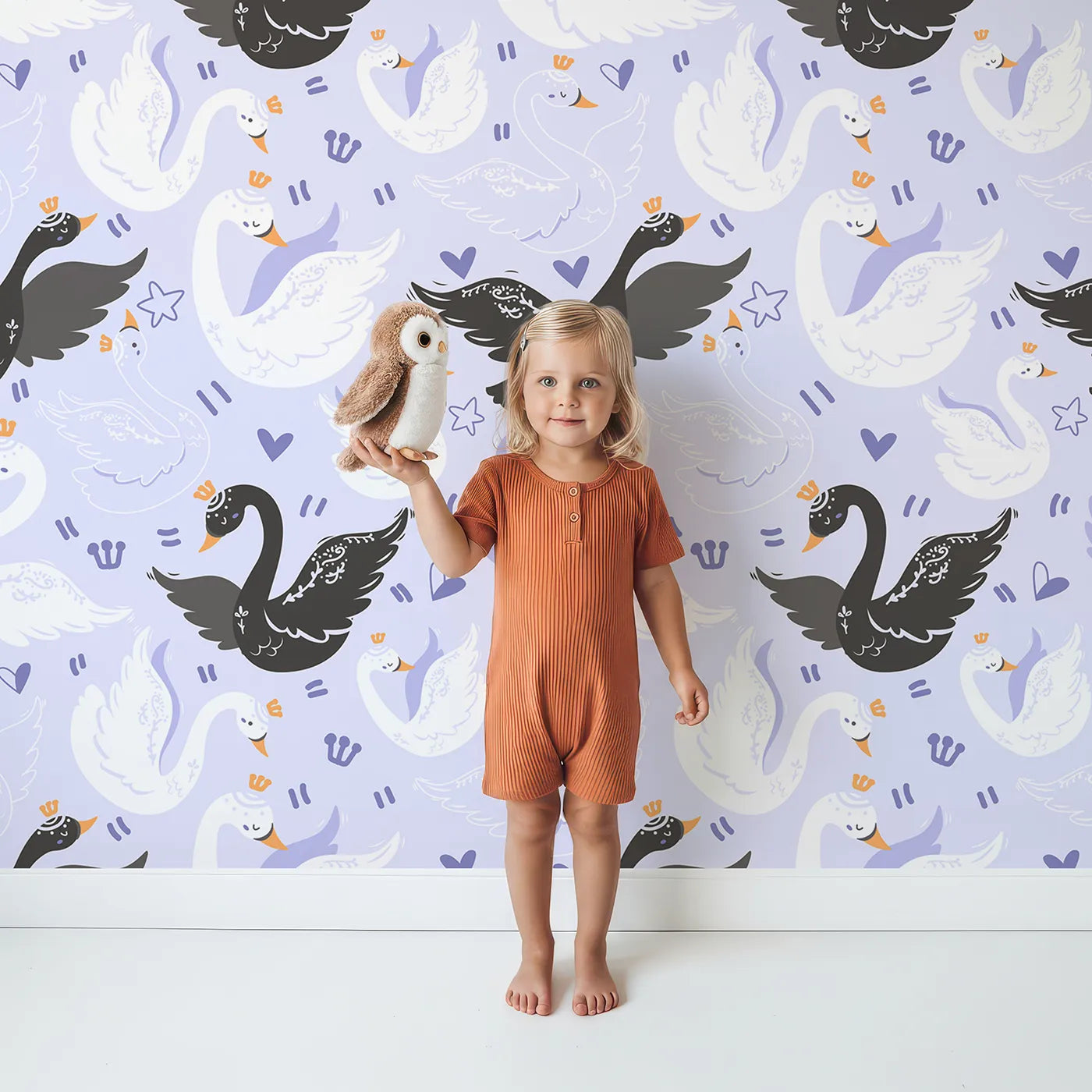 Lovely child standing in front of Whimsy Tots Royal Swan Parade Wallpaper in Lavender with swan, crown, heart, star, wing in design.