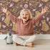 Lovely child standing in front of Whimsy Tots Floral Snuggle Bears Wallpaper in Berry Plum with bear, flower, heart, leaf in design. Lovely child standing in front of Whimsy Tots Floral Snuggle Bears Wallpaper in Berry Plum with bear, flower, heart, leaf in design.