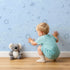 Lovely child standing in front of Whimsy Tots Floral Fox Cuddles Wallpaper in Sky Blue with fox, flower, leaf in design. Lovely child standing in front of Whimsy Tots Floral Fox Cuddles Wallpaper in Sky Blue with fox, flower, leaf in design.