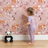 Lovely child standing in front of Whimsy Tots Folk Bloom Foxes Wallpaper in Rosewood Pink with fox, flower, vine in design. Lovely child standing in front of Whimsy Tots Folk Bloom Foxes Wallpaper in Rosewood Pink with fox, flower, vine in design.