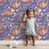 Lovely child standing in front of Whimsy Tots Folk Bloom Foxes Wallpaper in Wild Violet with fox, flower, vine in design. Lovely child standing in front of Whimsy Tots Folk Bloom Foxes Wallpaper in Wild Violet with fox, flower, vine in design.