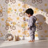 Lovely child standing in front of Whimsy Tots Honeybee Gnome Parade Wallpaper in Buttercream with gnome, bee, flower, bicycle in design. Lovely child standing in front of Whimsy Tots Honeybee Gnome Parade Wallpaper in Buttercream with gnome, bee, flower, bicycle in design.