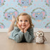 Lovely child standing in front of Whimsy Tots Floral Elephant Wink Wallpaper in Sky Blue with elephant, heart, flower, crown, branch, leaf in design. Lovely child standing in front of Whimsy Tots Floral Elephant Wink Wallpaper in Sky Blue with elephant, heart, flower, crown, branch, leaf in design.