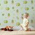 Lovely child standing in front of Whimsy Tots Hatching Dino Friends Wallpaper in Mint Meadow Green with dinosaur, egg, leaf, flower, bone in design. Lovely child standing in front of Whimsy Tots Hatching Dino Friends Wallpaper in Mint Meadow Green with dinosaur, egg, leaf, flower, bone in design.