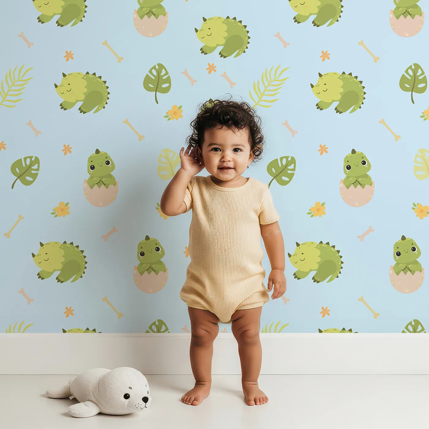 Lovely child standing in front of Whimsy Tots Hatching Dino Friends Wallpaper in Sky Blue with dinosaur, egg, leaf, flower, bone in design.