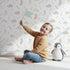 Lovely child standing in front of Whimsy Tots Fossil Roar Parade Wallpaper in Soft Taupe Beige with dinosaur, skeleton, bone, footprint in design. Lovely child standing in front of Whimsy Tots Fossil Roar Parade Wallpaper in Soft Taupe Beige with dinosaur, skeleton, bone, footprint in design.