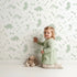 Lovely child standing in front of Whimsy Tots Dino Fossil Footprints Wallpaper in Sage Green with dinosaur, fossil, bone, footprint in design. Lovely child standing in front of Whimsy Tots Dino Fossil Footprints Wallpaper in Sage Green with dinosaur, fossil, bone, footprint in design.