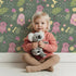 Lovely child standing in front of Whimsy Tots Cuddle Saurus Garden Wallpaper in Forest Fern Green with dinosaur, flower, bone, footprint in design. Lovely child standing in front of Whimsy Tots Cuddle Saurus Garden Wallpaper in Forest Fern Green with dinosaur, flower, bone, footprint in design.