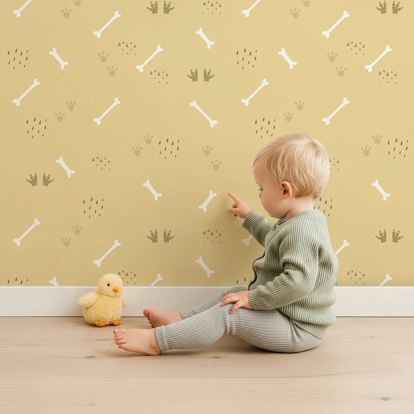 Lovely child standing in front of Whimsy Tots Dino Dig Basics Wallpaper in Fossil Beige with bone, footprint, raindrop, track in design.