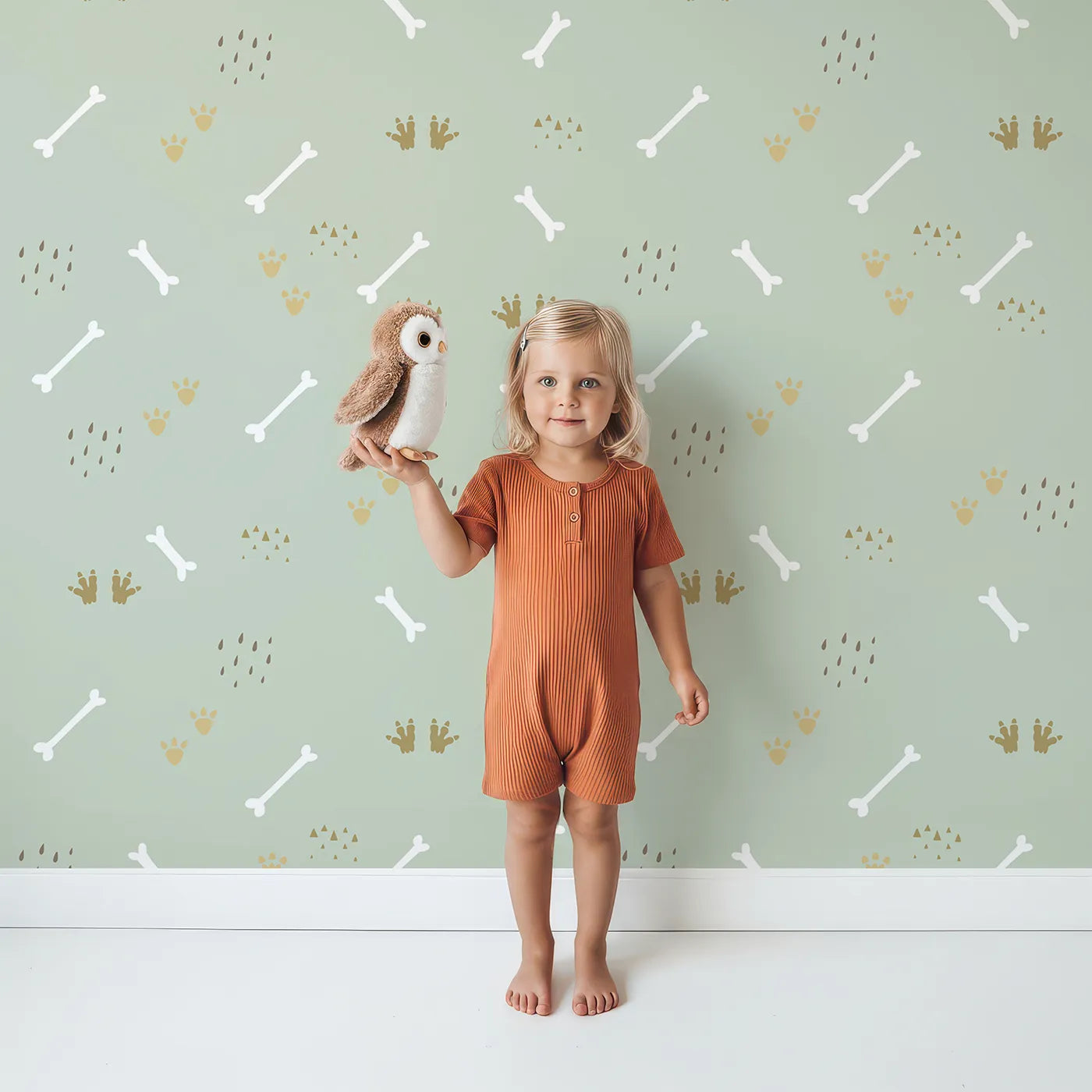 Lovely child standing in front of Whimsy Tots Dino Dig Basics Wallpaper in Sage Grove Green with bone, footprint, raindrop, track in design.