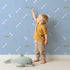 Lovely child standing in front of Whimsy Tots Dino Dig Basics Wallpaper in Dusk Blue with bone, footprint, raindrop, track in design. Lovely child standing in front of Whimsy Tots Dino Dig Basics Wallpaper in Dusk Blue with bone, footprint, raindrop, track in design.
