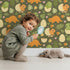 Lovely child standing in front of Whimsy Tots Hatchling Dino Garden Wallpaper in Olive Fern Green with dinosaur, egg, flower, leaf in design. Lovely child standing in front of Whimsy Tots Hatchling Dino Garden Wallpaper in Olive Fern Green with dinosaur, egg, flower, leaf in design.