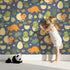 Lovely child standing in front of Whimsy Tots Hatchling Dino Garden Wallpaper in Midnight Nest with dinosaur, egg, flower, leaf in design. Lovely child standing in front of Whimsy Tots Hatchling Dino Garden Wallpaper in Midnight Nest with dinosaur, egg, flower, leaf in design.