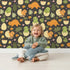 Lovely child standing in front of Whimsy Tots Hatchling Dino Garden Wallpaper in Charcoal Nest Gray with dinosaur, egg, flower, leaf in design. Lovely child standing in front of Whimsy Tots Hatchling Dino Garden Wallpaper in Charcoal Nest Gray with dinosaur, egg, flower, leaf in design.