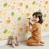 Lovely child standing in front of Whimsy Tots Hatchling Dino Parade Wallpaper in Dappled Beige with dinosaur, egg, bone, flower in design. Lovely child standing in front of Whimsy Tots Hatchling Dino Parade Wallpaper in Dappled Beige with dinosaur, egg, bone, flower in design.