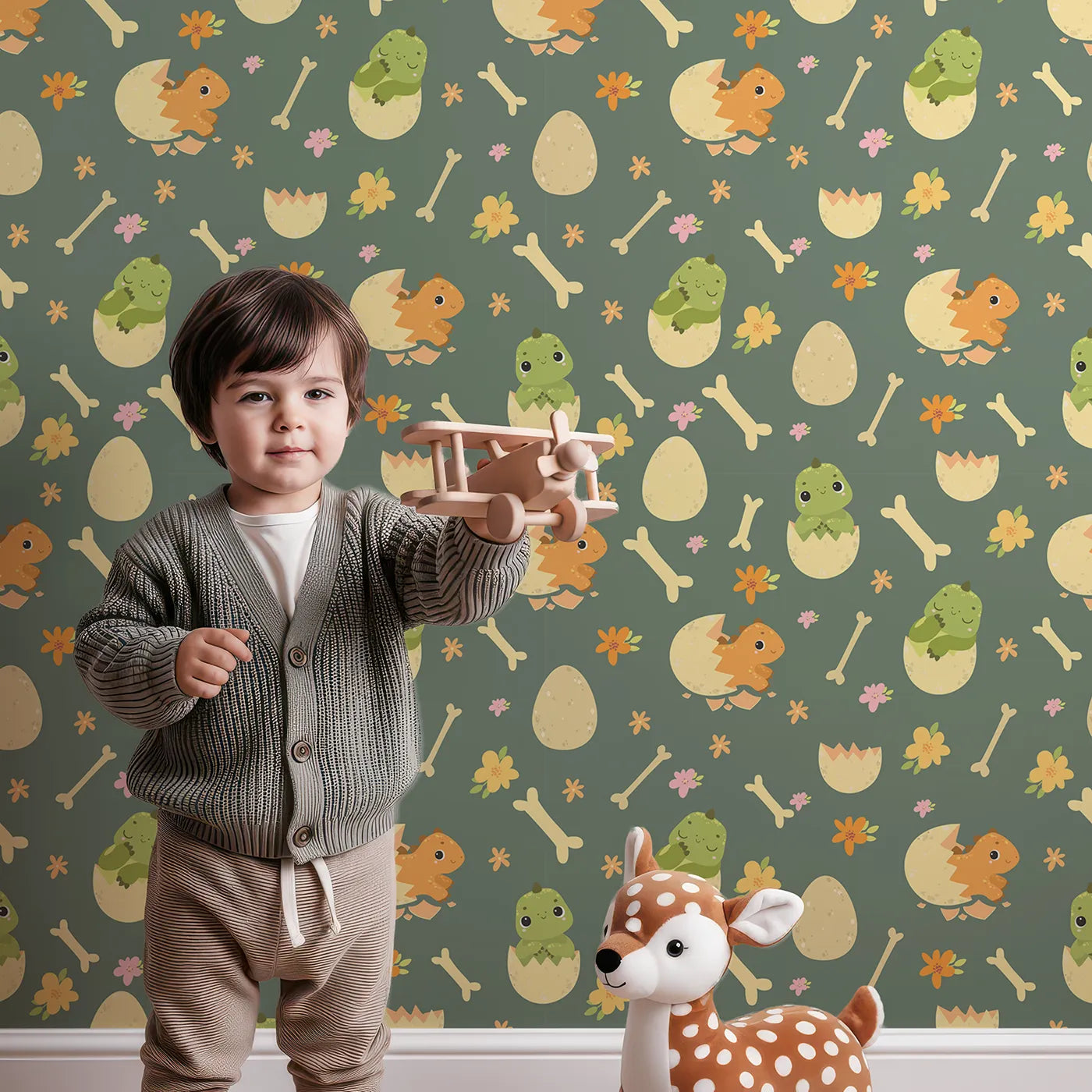 Lovely child standing in front of Whimsy Tots Hatchling Dino Parade Wallpaper in Olive Grove Green with dinosaur, egg, bone, flower in design.