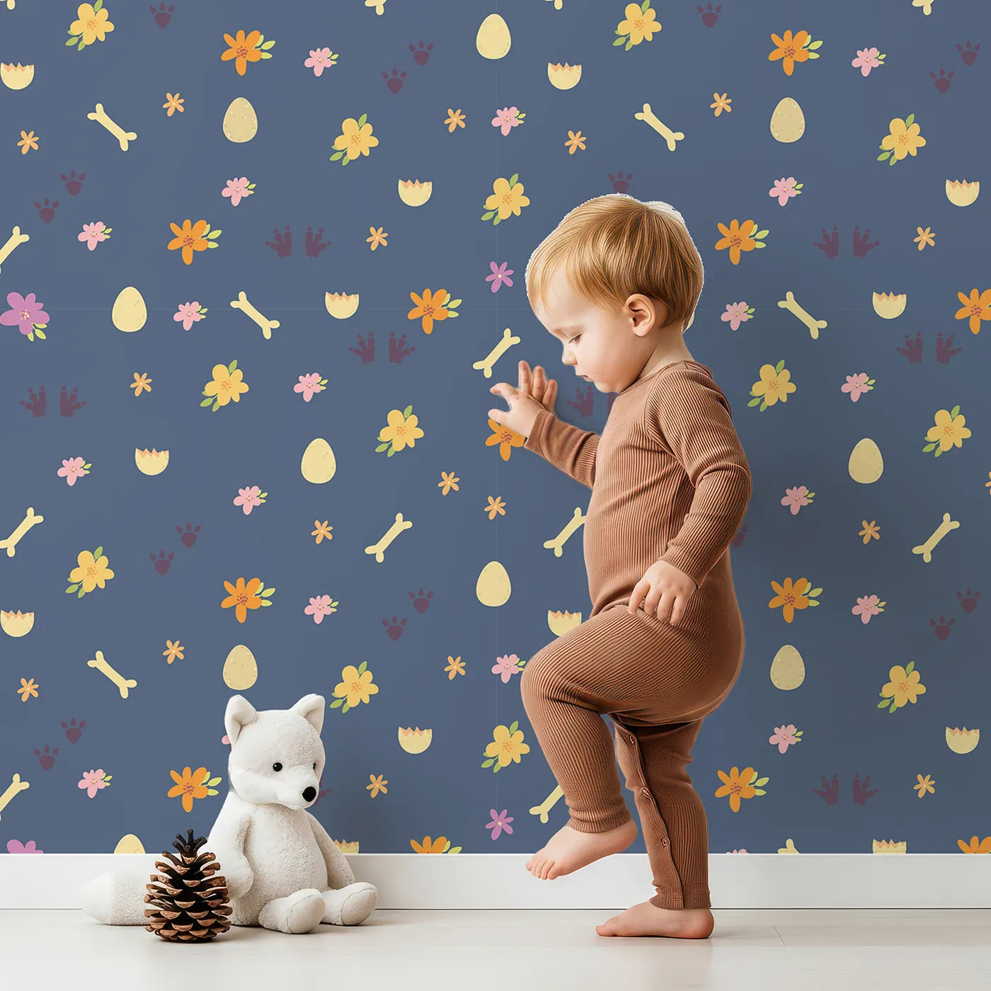 Lovely child standing in front of Whimsy Tots Dino Hatch Trail Wallpaper in Dusty Denim with egg, bone, flower, footprint in design.