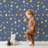 Lovely child standing in front of Whimsy Tots Dino Hatch Trail Wallpaper in Dusty Denim with egg, bone, flower, footprint in design. Lovely child standing in front of Whimsy Tots Dino Hatch Trail Wallpaper in Dusty Denim with egg, bone, flower, footprint in design.