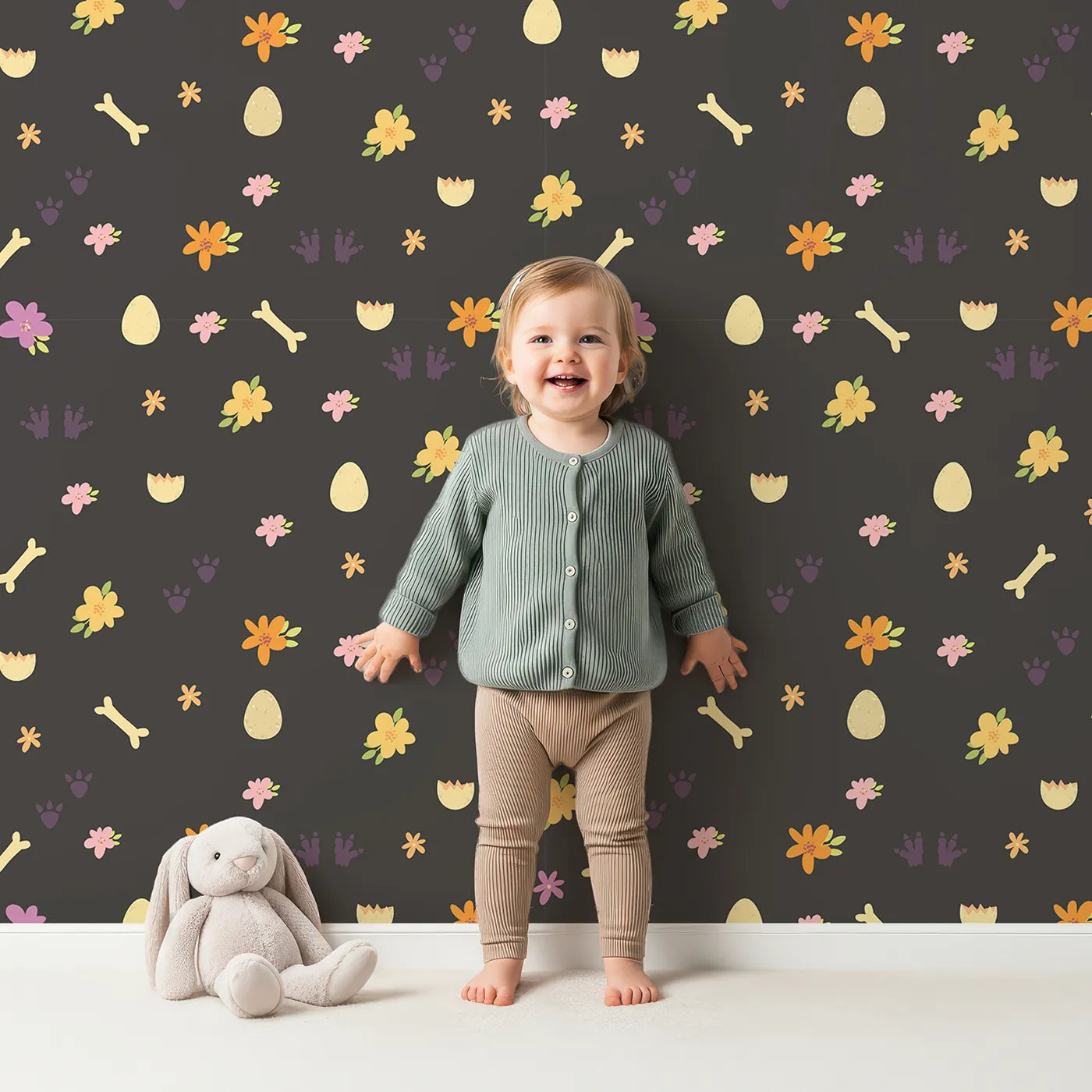 Lovely child standing in front of Whimsy Tots Dino Hatch Trail Wallpaper in Midnight Soil with egg, bone, flower, footprint in design.
