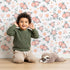 Lovely child standing in front of Whimsy Tots Whispering Peony Garden Wallpaper in Petal White with flower, peony, daisy, leaf in design. Lovely child standing in front of Whimsy Tots Whispering Peony Garden Wallpaper in Petal White with flower, peony, daisy, leaf in design.