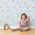 Lovely child standing in front of Whimsy Tots Floating Botanical Whispers Wallpaper in Morning Sky with flower, leaf, feather, peony in design. Lovely child standing in front of Whimsy Tots Floating Botanical Whispers Wallpaper in Morning Sky with flower, leaf, feather, peony in design.