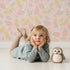 Lovely child standing in front of Whimsy Tots Floral Feather Drift Wallpaper in Blush Bloom Pink with feather, flower, blossom, leaf in design. Lovely child standing in front of Whimsy Tots Floral Feather Drift Wallpaper in Blush Bloom Pink with feather, flower, blossom, leaf in design.