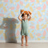 Lovely child standing in front of Whimsy Tots Floral Feather Drift Wallpaper in Sky Bloom with feather, flower, blossom, leaf in design. Lovely child standing in front of Whimsy Tots Floral Feather Drift Wallpaper in Sky Bloom with feather, flower, blossom, leaf in design.