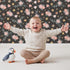 Lovely child standing in front of Whimsy Tots Twilight Garden Whimsy Wallpaper in Midnight Petal with flower, leaf, star, bloom in design. Lovely child standing in front of Whimsy Tots Twilight Garden Whimsy Wallpaper in Midnight Petal with flower, leaf, star, bloom in design.