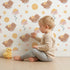 Lovely child standing in front of Whimsy Tots Moonlit Bear Dreams Wallpaper in Cream Sky with bear, flower, moon, star in design. Lovely child standing in front of Whimsy Tots Moonlit Bear Dreams Wallpaper in Cream Sky with bear, flower, moon, star in design.